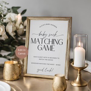May include: A gold-framed sign with white background and black text. The sign reads "Alicia's Baby Shower baby sock MATCHING GAME EACH GUEST GETS 10 SECONDS TO MATCH THE SOCKS WHOEVER MATCHES THE MOST - WINS! good luck!"