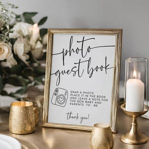 May include: A gold framed sign with the words "photo guest book" in elegant script. Below, a camera graphic and instructions to leave a note for the new baby and parents. A candle in a gold holder and a gold votive are also visible.