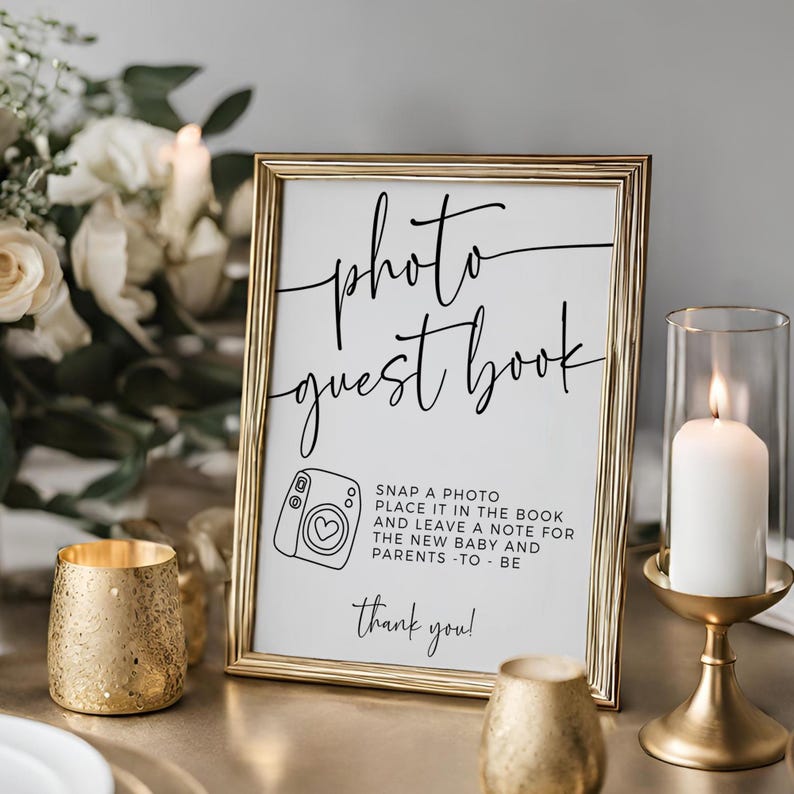 May include: A gold framed sign with the words "photo guest book" in elegant script. Below, a camera graphic and instructions to leave a note for the new baby and parents. A candle in a gold holder and a gold votive are also visible.
