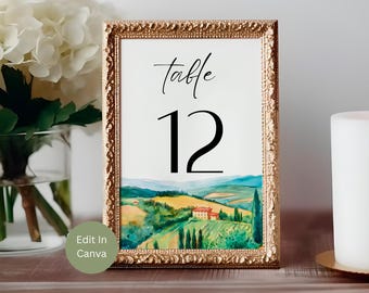Puede incluir: Un número de mesa enmarcado, el 12, con un marco dorado ornamentado. La obra de arte interior representa un paisaje de acuarela de colinas, campos y una casa. La palabra "mesa" está escrita encima del número.