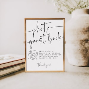 May include: A framed sign with the words "photo guest book" in elegant script. Below, a camera illustration and text that reads "Snap a photo, place it in the book and leave a note for the new baby and parents - to - be. Thank you!"