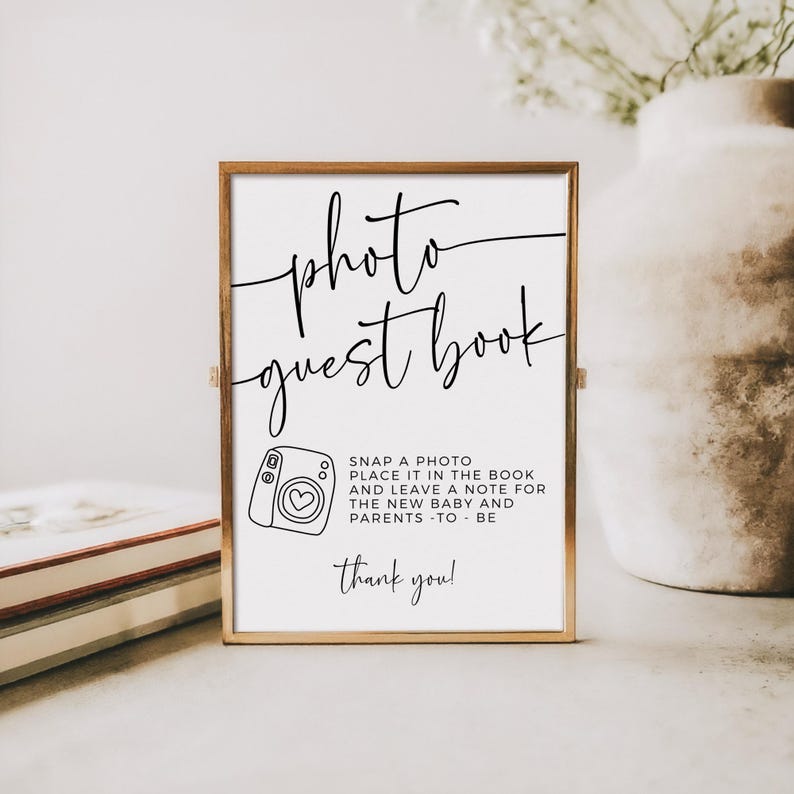 May include: A framed sign with the words "photo guest book" in elegant script. Below, a camera illustration and text that reads "Snap a photo, place it in the book and leave a note for the new baby and parents - to - be. Thank you!"