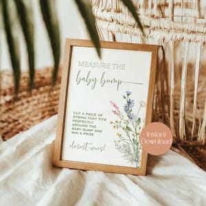 May include: A wooden framed sign with the text "Measure the baby bump" and instructions for a baby shower game. The sign is decorated with floral illustrations in pink, blue, and yellow. A circular sticker with the words "Instant Download" is on the bottom right.