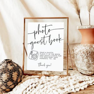 May include: A framed sign with the words "photo guest book" in elegant script. Below, it reads "Snap a photo, place it in the book and leave a note for the new baby and parents - to be. Thank you!" A camera icon is also present.