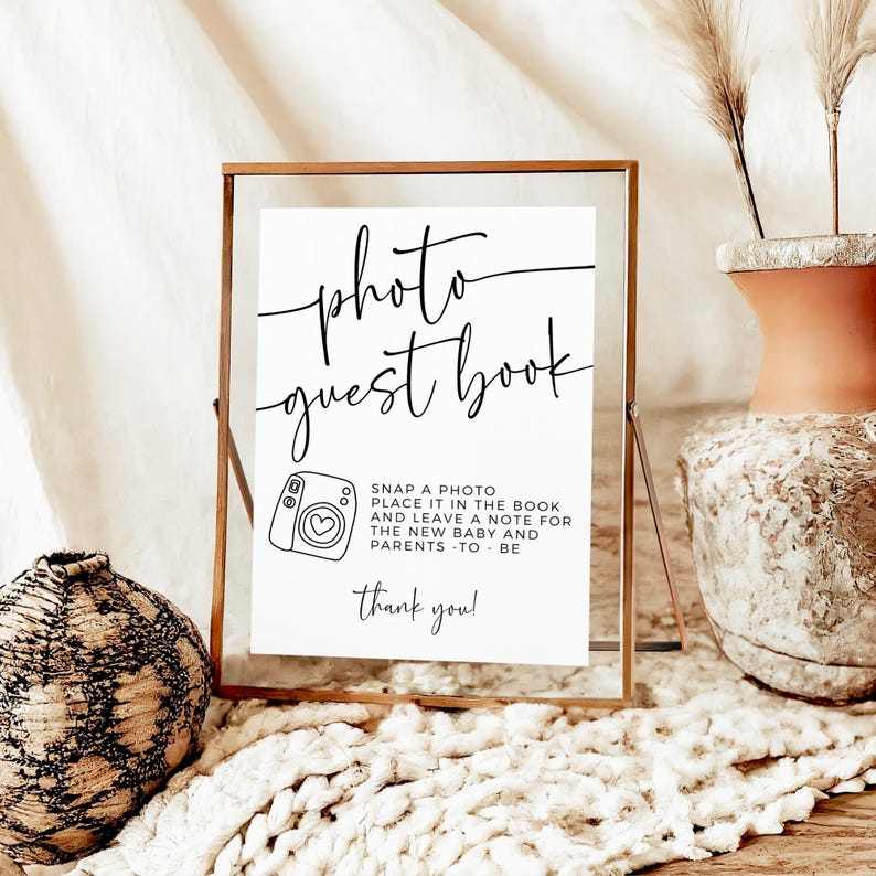May include: A framed sign with the words "photo guest book" in elegant script. Below, it reads "Snap a photo, place it in the book and leave a note for the new baby and parents - to be. Thank you!" A camera icon is also present.