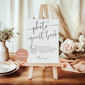 May include: A white sign on a small wooden easel with the words "photo guest book" in black script. Below is a camera graphic and text that reads "Snap a photo, place it in the book and leave a note for the new baby and parents - to be. Thank you!"