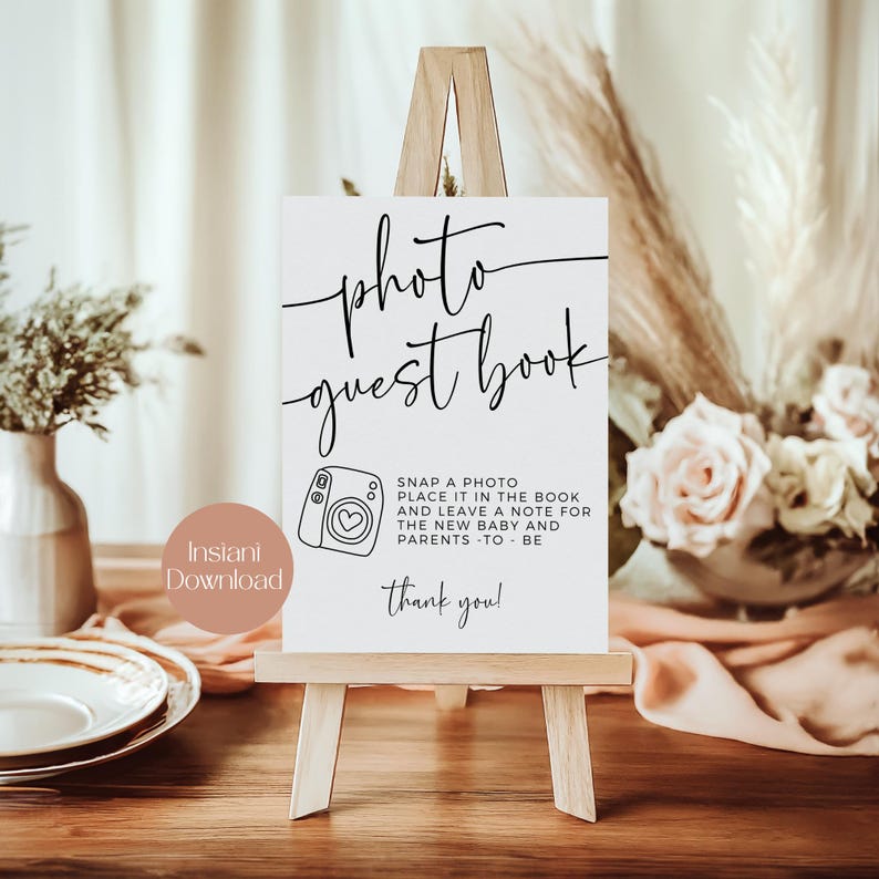 May include: A white sign on a small wooden easel with the words "photo guest book" in black script. Below is a camera graphic and text that reads "Snap a photo, place it in the book and leave a note for the new baby and parents - to be. Thank you!"
