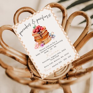 May include: A baby shower invitation with a watercolor illustration of pancakes topped with berries and syrup, and a pacifier. The text reads "pancakes & pacifiers" and includes event details: date, time, location, and RSVP information.