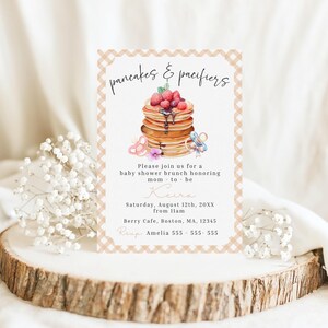 May include: A baby shower invitation featuring a stack of pancakes topped with berries and syrup, alongside a pacifier and flowers. The text reads "pancakes & pacifiers" and includes event details: date, time, location, and RSVP information.
