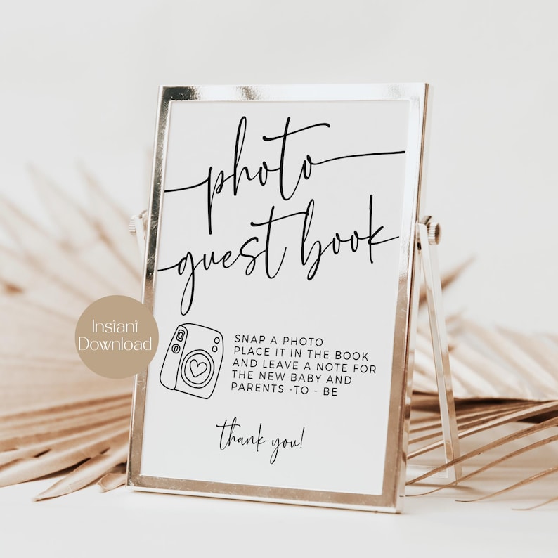 May include: A framed sign with the words "photo guest book" in elegant script. The sign includes instructions to snap a photo and leave a note. A small camera graphic is also present. The sign is in a silver frame.