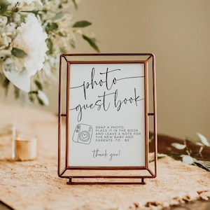 May include: A framed sign with the words "photo guest book" in elegant script. The sign includes instructions to snap a photo and leave a note for the new baby and parents. The frame is a warm copper color, and the background is neutral.