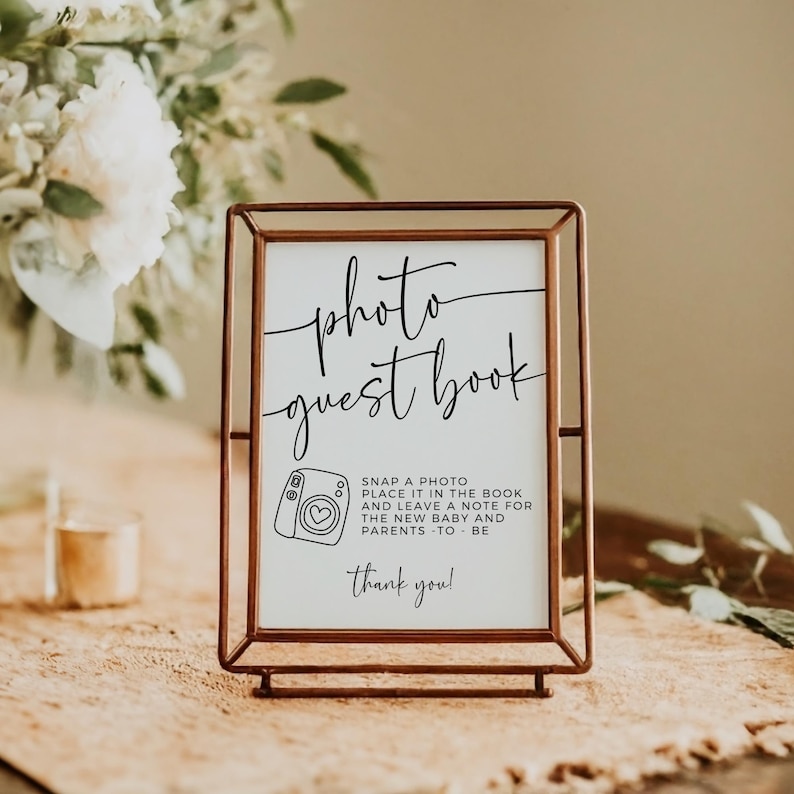 May include: A framed sign with the words "photo guest book" in elegant script. The sign includes instructions to snap a photo and leave a note for the new baby and parents. The frame is a warm copper color, and the background is neutral.