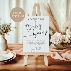 May include: White sign with black text on an easel. The sign says "Measure the Baby Bump. Cut a piece of string that fits perfectly around the baby bump and win a prize. Good luck!"