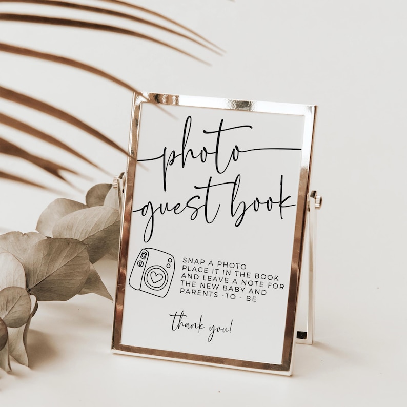 May include: A framed sign with the words "photo guest book" in elegant script. The sign includes instructions to snap a photo and leave a note for the new baby and parents-to-be, with a thank you message at the bottom. A camera illustration is also included.