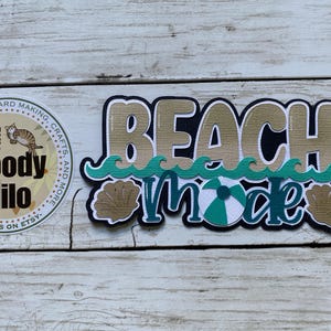 May include: A decorative sign with the words "BEACH mode" in a layered design. The sign features a beach ball, seashells, and wave accents in teal, tan, and black. A round sticker with the text "Moody Milo" is also visible.