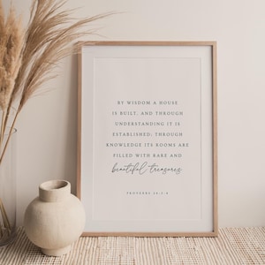 May include: A framed print with the text "By wisdom a house is built...beautiful treasures" in a minimalist style. The print is in a light wood frame. A vase with dried pampas grass and a ceramic vase are on a woven surface.