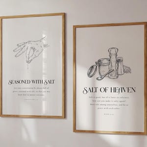 以下が含まれることがあります： ミニマリストデザインの額入りアートプリント2枚。片方は「SEASONED WITH SALT」の文字とともに塩を振りかける手を描いています。もう片方は「SALT OF HEAVEN」の文字とともに塩に関連するアイテムを描いています。