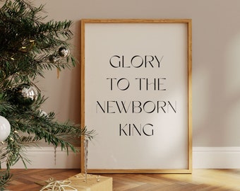 May include: A framed print with the text "Glory to the Newborn King" in black lettering on a white background. The frame is made of natural wood and has a light brown finish. The print is sitting on a wooden floor with a Christmas tree in the background.