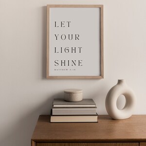 May include: A framed print with the text "LET YOUR LIGHT SHINE" and the scripture reference "MATTHEW 5:16". The print is on a wooden shelf with a stack of books and a white ceramic vase.