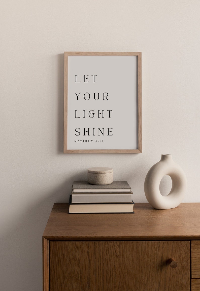 May include: A framed print with the text "LET YOUR LIGHT SHINE" and the scripture reference "MATTHEW 5:16". The print is on a wooden shelf with a stack of books and a white ceramic vase.