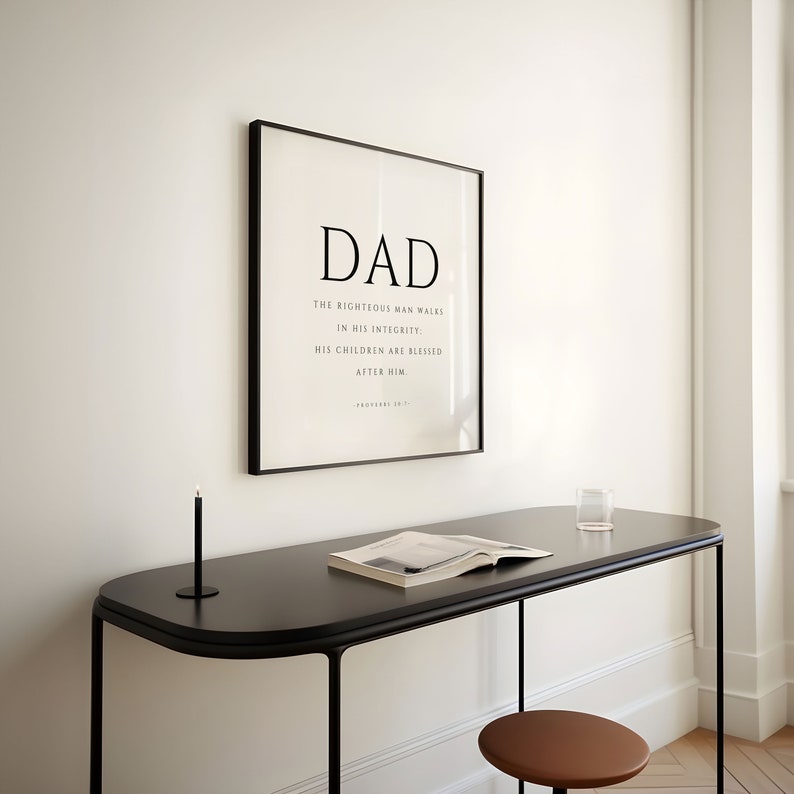 Puede incluir: Impresi&oacute;n enmarcada en blanco y negro con la palabra "DAD" y una cita sobre un hombre justo que camina en su integridad. La cita es de Proverbios 20:7. La impresi&oacute;n est&aacute; colgada en una pared blanca sobre un escritorio negro con un libro y un vaso de agua encima.