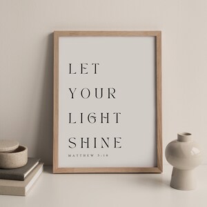 May include: A framed print with the text "Let your light shine" and the scripture reference "Matthew 5:16". The print is in a light wood frame and is sitting on a white surface.