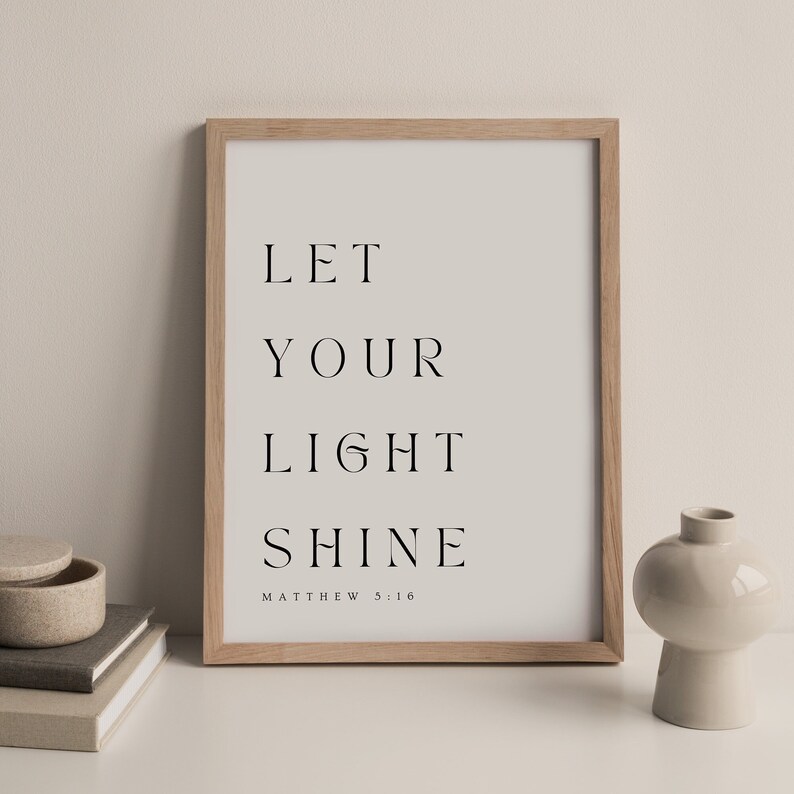 May include: A framed print with the text "Let your light shine" and the scripture reference "Matthew 5:16". The print is in a light wood frame and is sitting on a white surface.