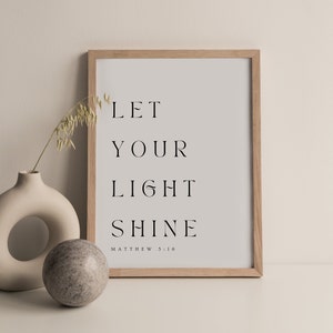 May include: A framed print with the text "Let your light shine" and the scripture reference "Matthew 5:16". The print is in a light wood frame and is sitting on a white surface.