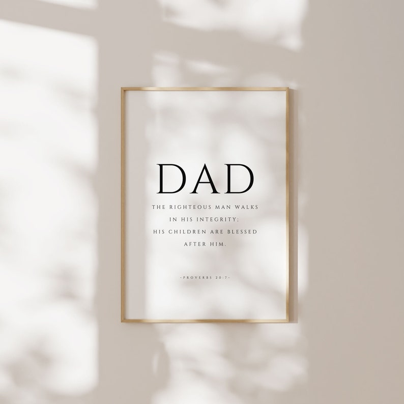 Puede incluir: Una impresi&oacute;n enmarcada con un marco dorado y un fondo blanco. La impresi&oacute;n presenta la palabra "DAD" en letras negras grandes, seguida del texto "El hombre justo camina en su integridad; sus hijos son bendecidos despu&eacute;s de &eacute;l." El texto est&aacute; en negro y se atribuye a "Proverbios 20:7".