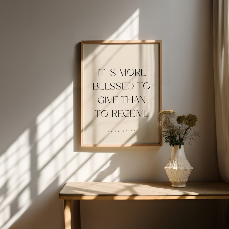 May include: A framed print with a beige background and black text that reads "It is more blessed to give than to receive. Acts 20:35". The print is hanging on a light beige wall with a wooden table and a white vase with flowers in front of it.
