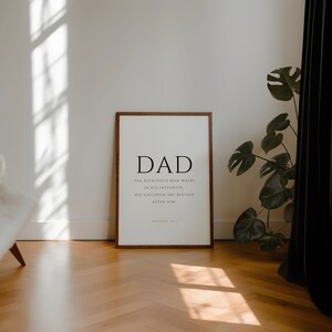 Puede incluir: Una impresi&oacute;n enmarcada con un marco de madera marr&oacute;n. La impresi&oacute;n es blanca con texto negro que dice "DAD" y una cita de Proverbios 20:7: "El hombre justo anda en su integridad: Sus hijos son bendecidos despu&eacute;s de &eacute;l."