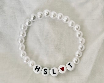 Peut inclure: Bracelet de perles blanches avec les lettres HSL et T en noir sur des perles blanches. Un cœur rouge remplace la lettre O. Le bracelet est composé de perles blanches et rondes.