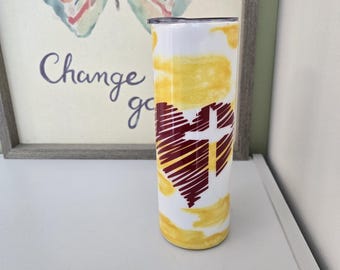 May include: A white, insulated tumbler with a yellow and white watercolor design. A maroon heart with a cross is featured on the front. The tumbler is cylindrical and has a black lid. A framed print with a butterfly and the words "Change go" is in the background.