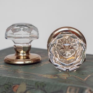 May include: Two clear glass door knobs with faceted designs and gold-coloured bases. One knob is upright, the other is on its side, showcasing the intricate starburst pattern. The knobs are on a green surface.