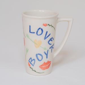 May include: White ceramic mug with a blue and green design. The mug has the words "Lover Boy" written on it in blue.