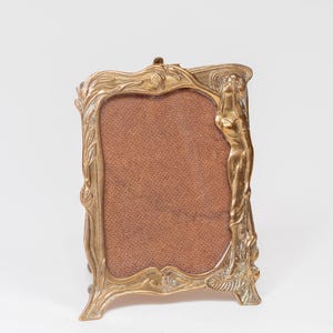 May include: An Art Nouveau style brass picture frame. The frame has a rectangular aperture with a brown backing. It features a standing female figure and floral detailing. The frame is supported by two small feet and is made of brass.