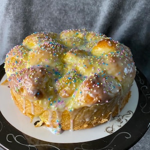 May include: A round loaf of bread with a white glaze and colorful sprinkles. The bread is on a white plate with a black and white design.