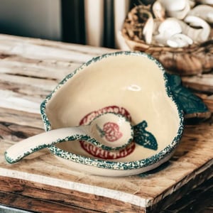 May include: A ceramic apple-shaped bowl with a matching spoon, both featuring a cream-colored base and a red apple design. The bowl and spoon have a green speckled trim. The bowl sits on a wooden surface.