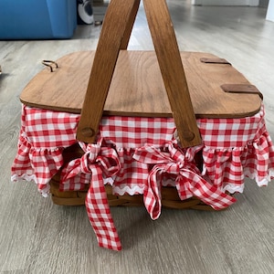 May include: A red and white gingham picnic basket with wooden handles and a wooden lid. The basket has a white lace trim and red bows.