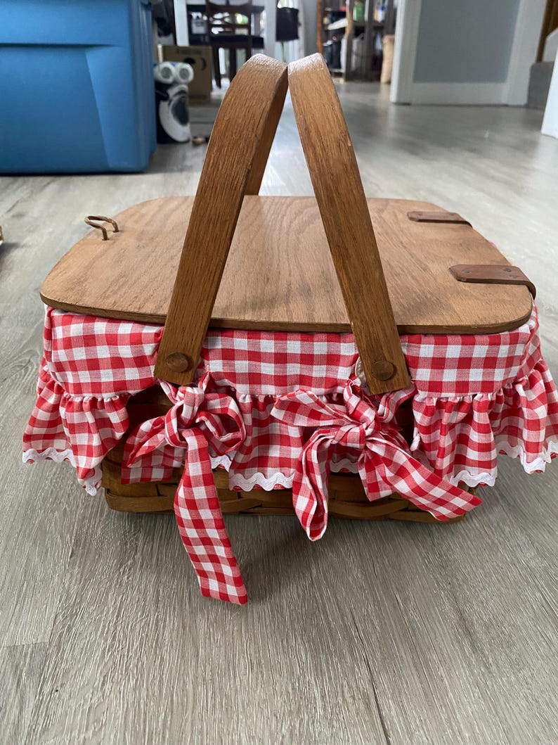 May include: A red and white gingham picnic basket with wooden handles and a wooden lid. The basket has a white lace trim and red bows.