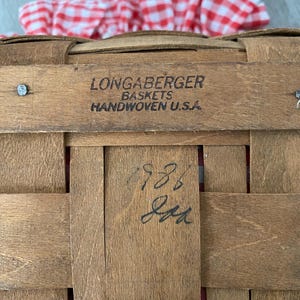 May include: A Longaberger basket with a red and white gingham cloth inside. The basket is made of woven wood and has a label that reads "Longaberger Baskets Handwoven U.S.A.". The basket also has a handwritten number "788" on the bottom.