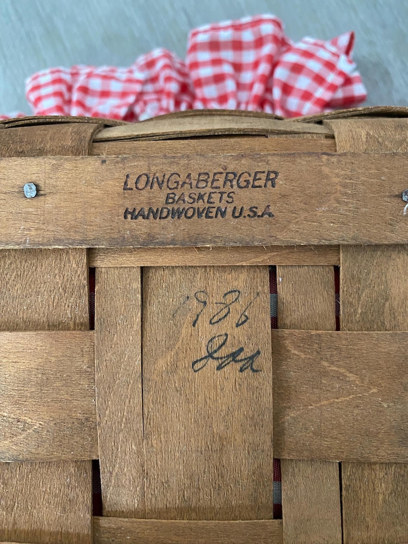 May include: A Longaberger basket with a red and white gingham cloth inside. The basket is made of woven wood and has a label that reads "Longaberger Baskets Handwoven U.S.A.". The basket also has a handwritten number "788" on the bottom.