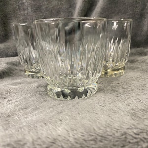 May include: Three clear glass tumblers with a cut glass design. The glasses are sitting on a gray fuzzy surface.