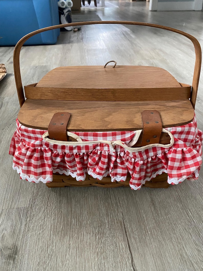 May include: A wooden picnic basket with a red and white gingham fabric lining and a brown leather handle.