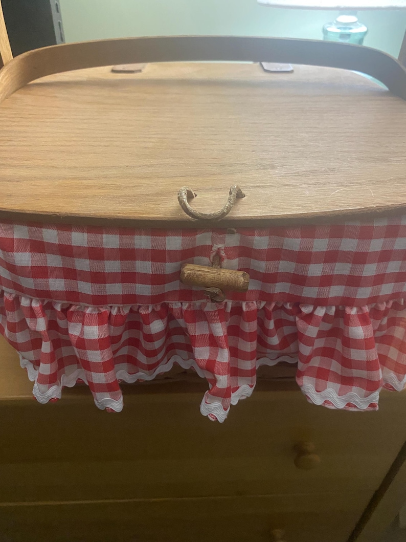 May include: A wooden picnic basket with a red and white gingham lining and a white lace trim. The basket has a wooden handle and a metal clasp.