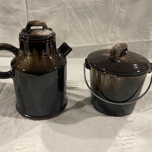 May include: A brown and white ceramic teapot and a matching creamer with a metal handle. The teapot has a lid and a spout. The creamer has a lid.