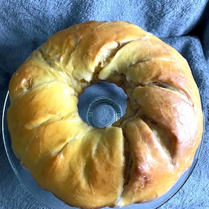May include: A golden-brown baked bundt cake with a glossy glaze, presented on a clear glass plate. The cake has a ring shape with a central hole, and is sitting on a clear glass plate.