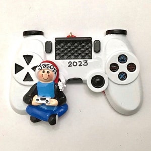 May include: White plastic video game controller ornament with a small figurine of a person wearing a Santa hat and holding a video game controller. The controller has black buttons and a black and white checkered design on the top. The year 2023 is printed on the controller.