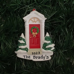 May include: A Christmas ornament featuring a red front door with a green wreath and a white frame. The ornament has two small snow-covered trees on either side of the door and the text "2023 The Brady's".