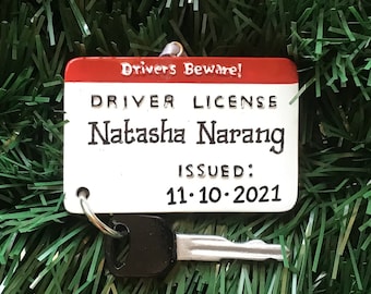 Può includere: Un portachiavi a forma di patente di guida. L'etichetta rossa e bianca recita "Drivers Beware!" e mostra il nome "Natasha Narang" e la data di emissione "11-10-2021". Una piccola chiave è attaccata al portachiavi.